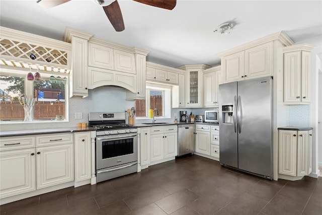 kitchen featuring cream cabinets, stainless steel appliances, dark countertops, ceiling fan, and a sink