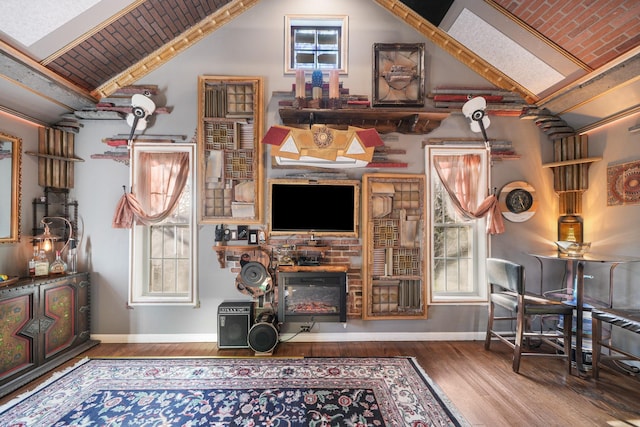 living area with vaulted ceiling, wood finished floors, baseboards, a glass covered fireplace, and brick ceiling