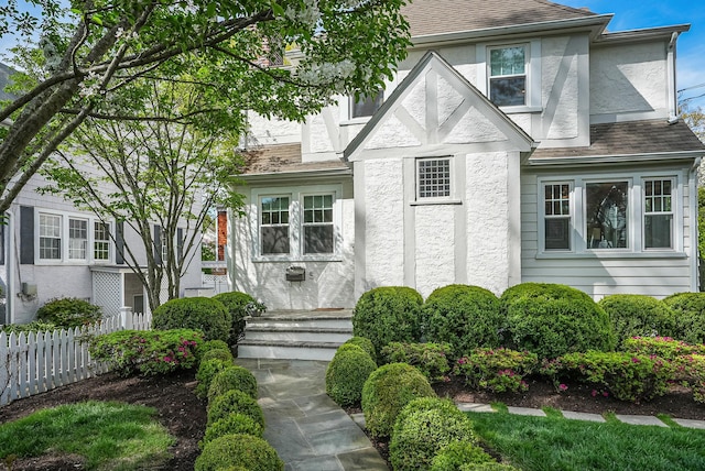 tudor-style house with fence, stucco siding, and a shingled roof