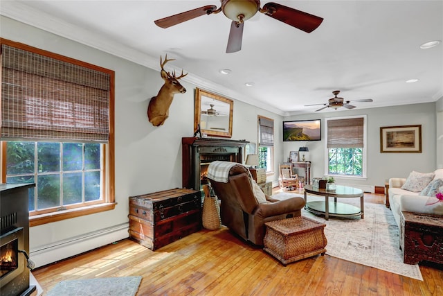 living room with a warm lit fireplace, baseboard heating, ceiling fan, crown molding, and hardwood / wood-style floors