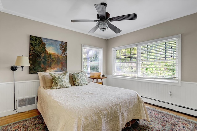 bedroom featuring a wainscoted wall, wood finished floors, a baseboard radiator, and a ceiling fan