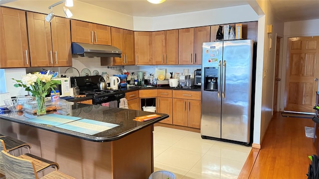 kitchen with black range with gas stovetop, under cabinet range hood, brown cabinetry, stainless steel refrigerator with ice dispenser, and a peninsula