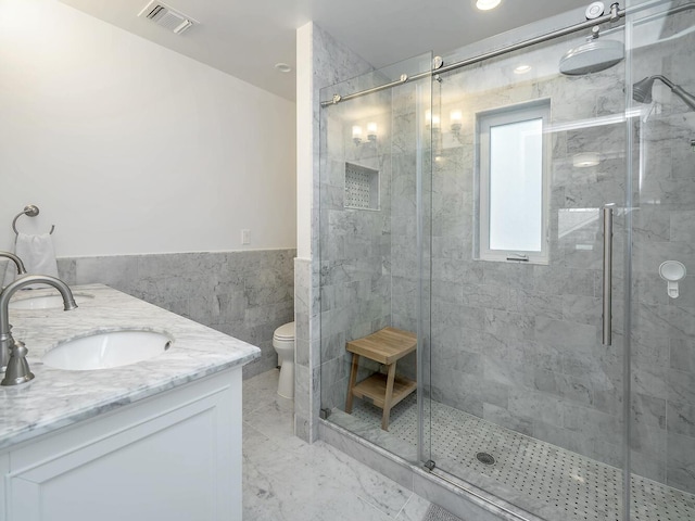 bathroom with tile walls, toilet, a shower stall, double vanity, and a wainscoted wall