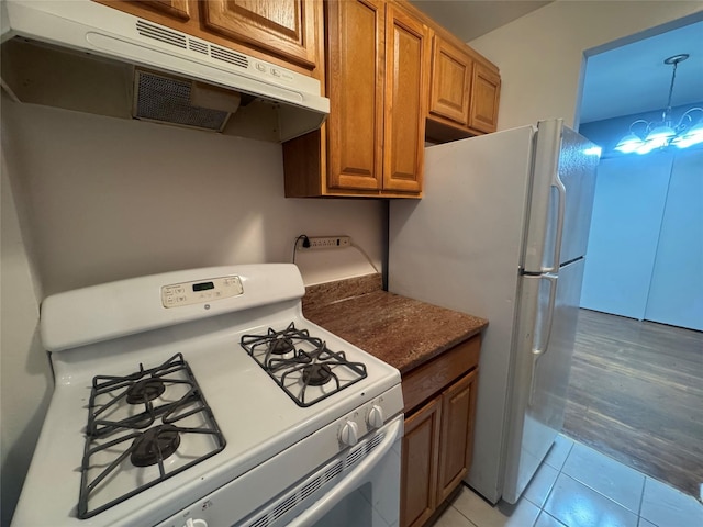 kitchen with white appliances, under cabinet range hood, brown cabinets, light tile patterned floors, and dark countertops