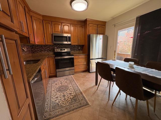 kitchen with dark stone counters, appliances with stainless steel finishes, brown cabinets, backsplash, and a sink