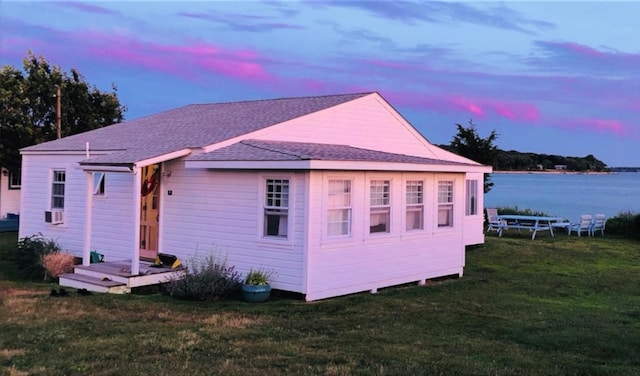 back of property at dusk with roof with shingles, a yard, and a water view