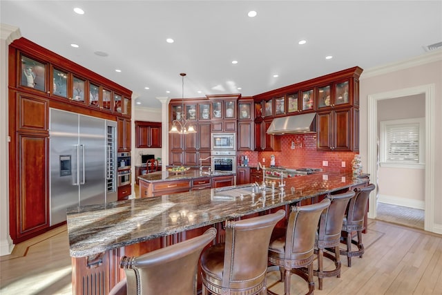kitchen with decorative backsplash, built in appliances, reddish brown cabinets, under cabinet range hood, and a sink