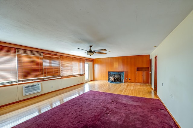unfurnished living room featuring wood finished floors, baseboards, ceiling fan, a wall unit AC, and a premium fireplace