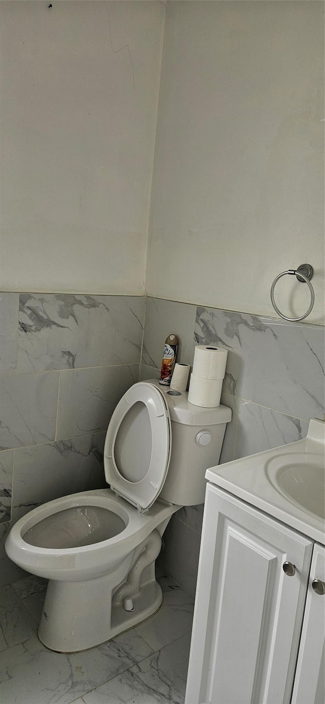 half bathroom with vanity, marble finish floor, toilet, and tile walls
