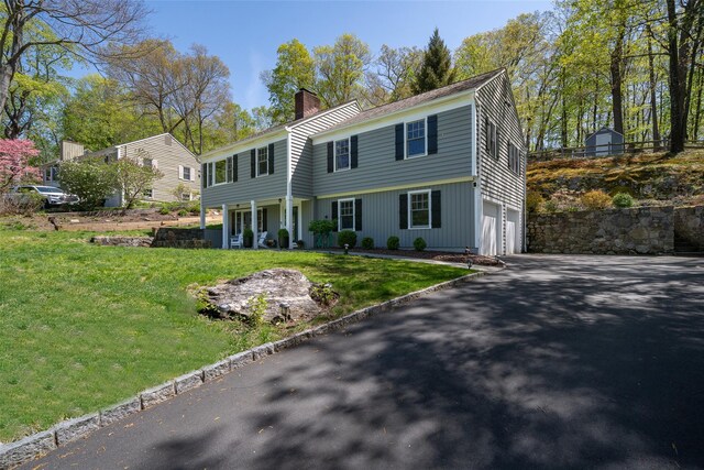colonial home with aphalt driveway, a garage, a front yard, a chimney, and covered porch