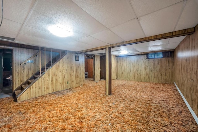 basement featuring a paneled ceiling, carpet flooring, wood walls, and stairs