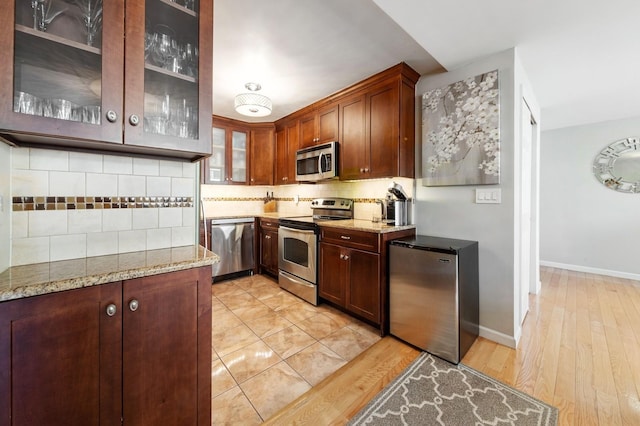 kitchen with light stone countertops, appliances with stainless steel finishes, light wood-style flooring, backsplash, and glass insert cabinets