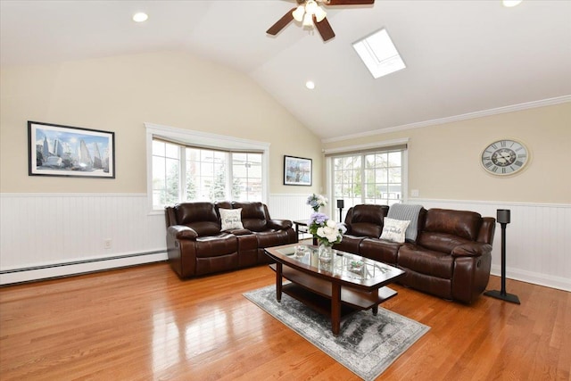 living area featuring vaulted ceiling with skylight, ceiling fan, a wainscoted wall, light wood-style flooring, and a baseboard radiator