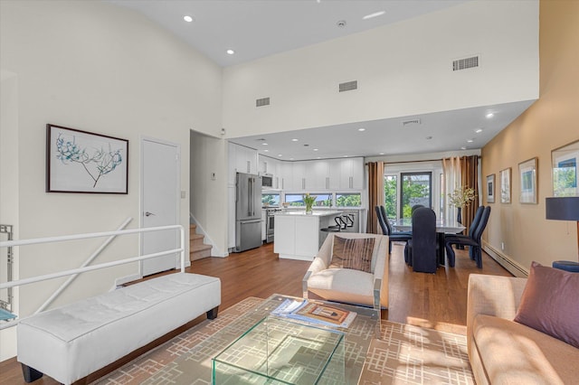 living room featuring a towering ceiling, a baseboard radiator, wood finished floors, and visible vents