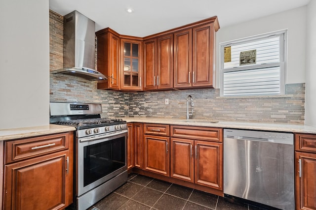 kitchen with appliances with stainless steel finishes, a sink, brown cabinetry, wall chimney exhaust hood, and glass insert cabinets