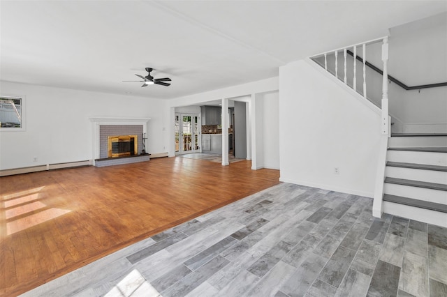 unfurnished living room with ceiling fan, a brick fireplace, wood finished floors, baseboards, and stairs