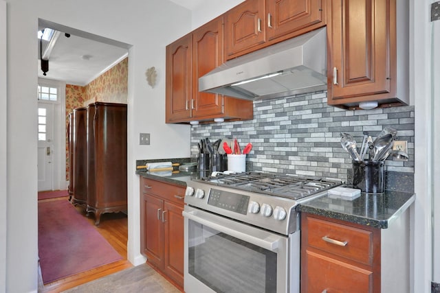 kitchen featuring decorative backsplash, brown cabinetry, gas range, dark stone countertops, and under cabinet range hood