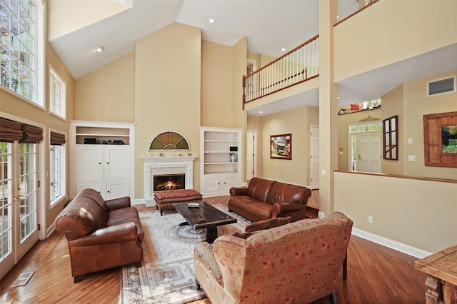 living area with a warm lit fireplace, baseboards, wood finished floors, a towering ceiling, and visible vents