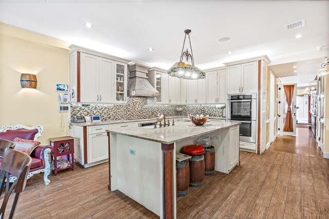 kitchen featuring backsplash, stainless steel double oven, visible vents, wood-type flooring, and custom range hood