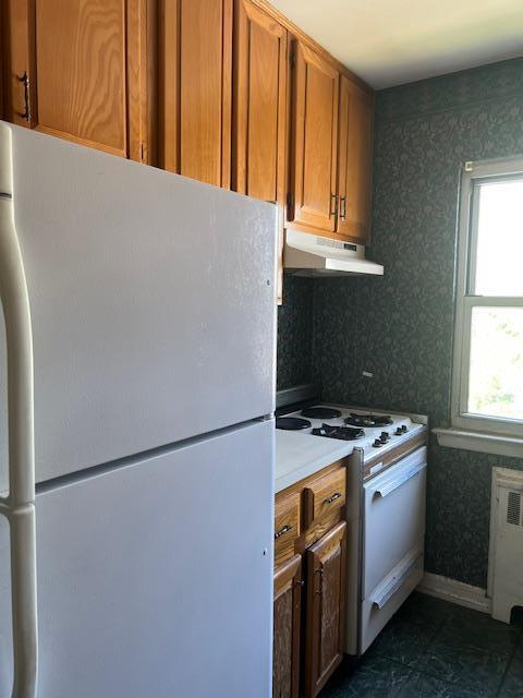 kitchen with brown cabinetry, radiator, under cabinet range hood, wallpapered walls, and white appliances