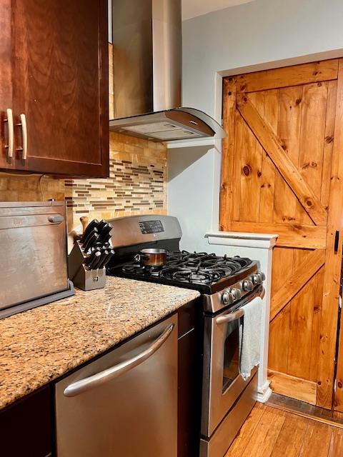 kitchen featuring wall chimney range hood, tasteful backsplash, light stone counters, light wood-style flooring, and appliances with stainless steel finishes