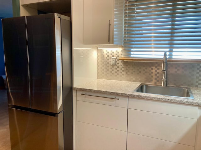 kitchen featuring freestanding refrigerator, white cabinets, light stone counters, a sink, and backsplash