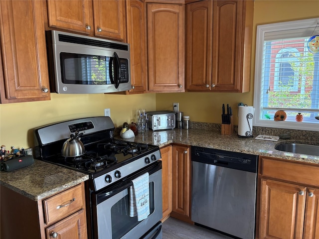 kitchen with dark stone counters, stainless steel appliances, brown cabinetry, and a sink