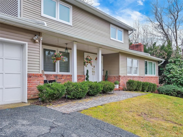 doorway to property featuring brick siding, a garage, a porch, a chimney, and a yard
