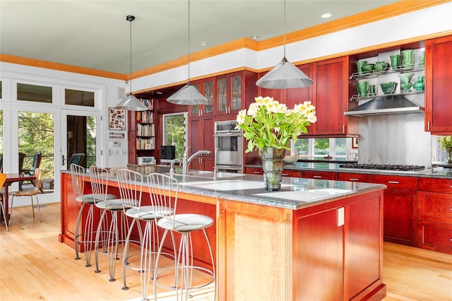 kitchen featuring a kitchen island with sink, dark brown cabinets, wall chimney exhaust hood, a sink, and stainless steel appliances