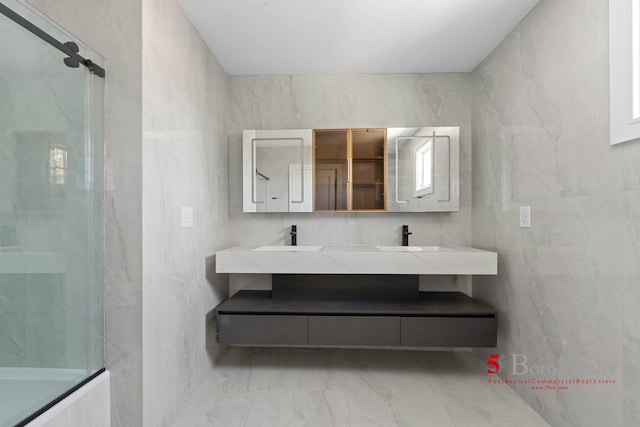 bathroom with a sink, a shower with shower door, double vanity, marble finish floor, and tile walls