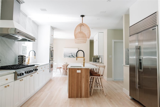 kitchen featuring wall chimney range hood, appliances with stainless steel finishes, light wood-style floors, white cabinetry, and an island with sink