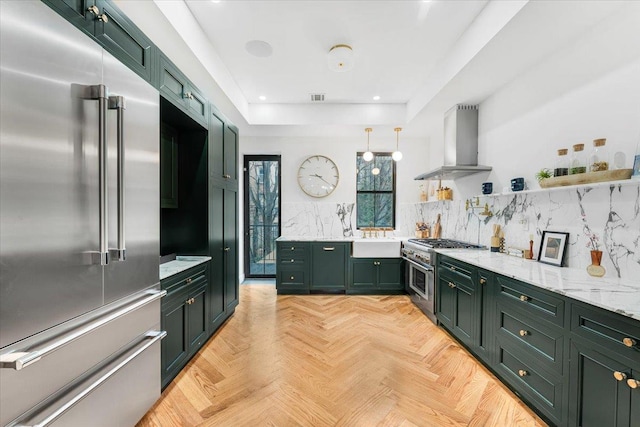 kitchen with decorative backsplash, high end appliances, wall chimney range hood, green cabinetry, and a tray ceiling