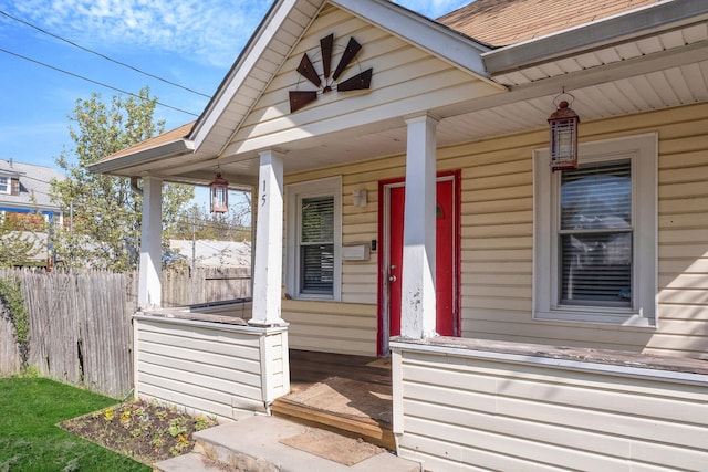 view of exterior entry with fence, covered porch, and roof with shingles