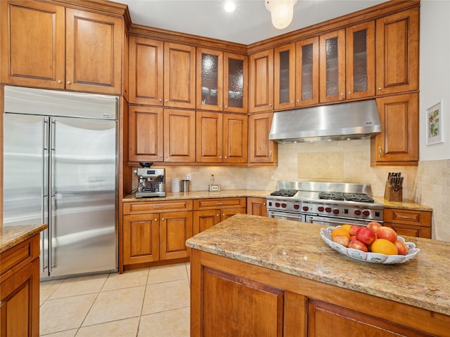 kitchen featuring under cabinet range hood, high quality appliances, light stone counters, and brown cabinetry