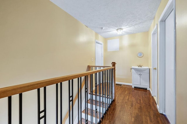 hall with a textured ceiling, wood finished floors, a sink, baseboards, and an upstairs landing