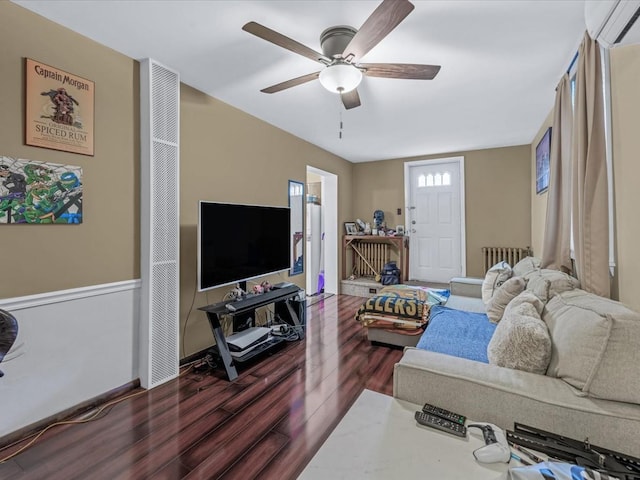 living area with wood finished floors, a wall unit AC, a ceiling fan, radiator heating unit, and baseboards