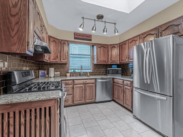 kitchen featuring stainless steel appliances, a sink, brown cabinetry, under cabinet range hood, and tasteful backsplash