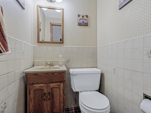 bathroom with wallpapered walls, wainscoting, toilet, vanity, and tile walls
