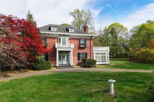 colonial-style house featuring brick siding, a front yard, a chimney, a balcony, and french doors
