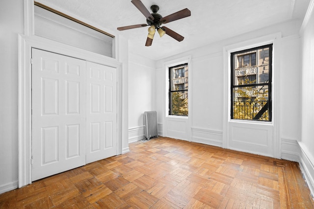 unfurnished bedroom featuring radiator heating unit, a decorative wall, a closet, ceiling fan, and wainscoting