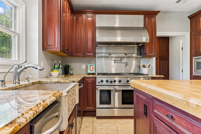 1465 Ackerly Pond Ln, Southold, NY 11971 - kitchen with wall chimney exhaust hood, stainless steel appliances, reddish brown cabinets, light tile patterned floors, and light stone counters
