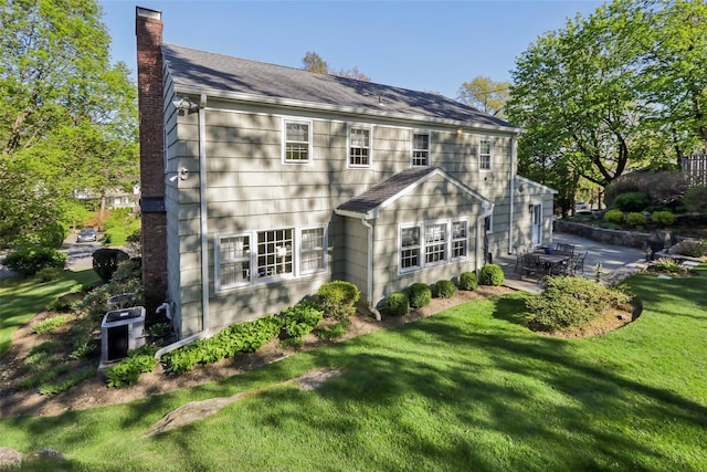 rear view of property with a chimney, a patio area, a lawn, and roof with shingles
