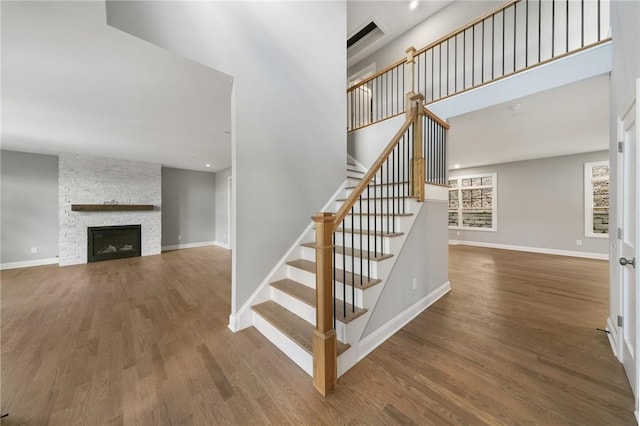 staircase featuring baseboards, a towering ceiling, wood finished floors, and a stone fireplace