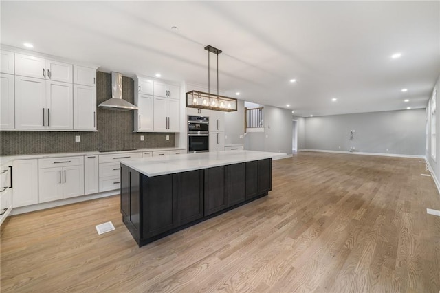 kitchen with wall chimney range hood, stainless steel double oven, light countertops, a center island, and white cabinetry