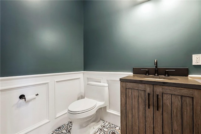 half bathroom with vanity, a decorative wall, toilet, tile patterned floors, and a wainscoted wall