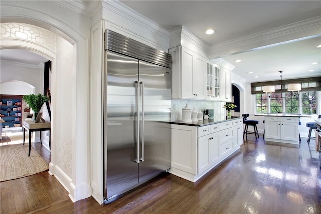 kitchen with stainless steel built in refrigerator, dark wood-type flooring, arched walkways, white cabinets, and crown molding