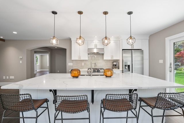 kitchen with stainless steel fridge, arched walkways, decorative backsplash, under cabinet range hood, and a spacious island