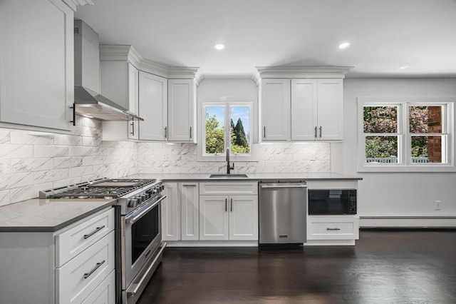 kitchen with a baseboard radiator, wall chimney exhaust hood, a sink, appliances with stainless steel finishes, and a wealth of natural light