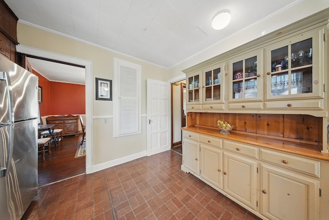 kitchen with crown molding, freestanding refrigerator, brick patterned floor, glass insert cabinets, and cream cabinetry