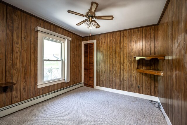 carpeted empty room featuring wooden walls, baseboard heating, a ceiling fan, baseboards, and ornamental molding